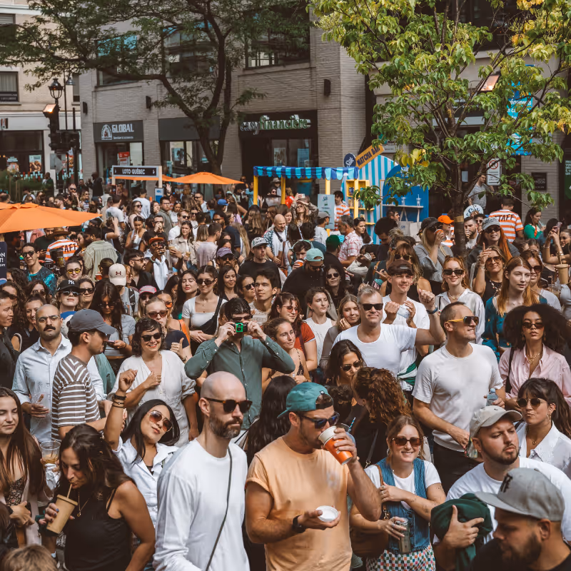 Large crowd of diverse people enjoying an outdoor event on a city street with trees and vendor stalls.
