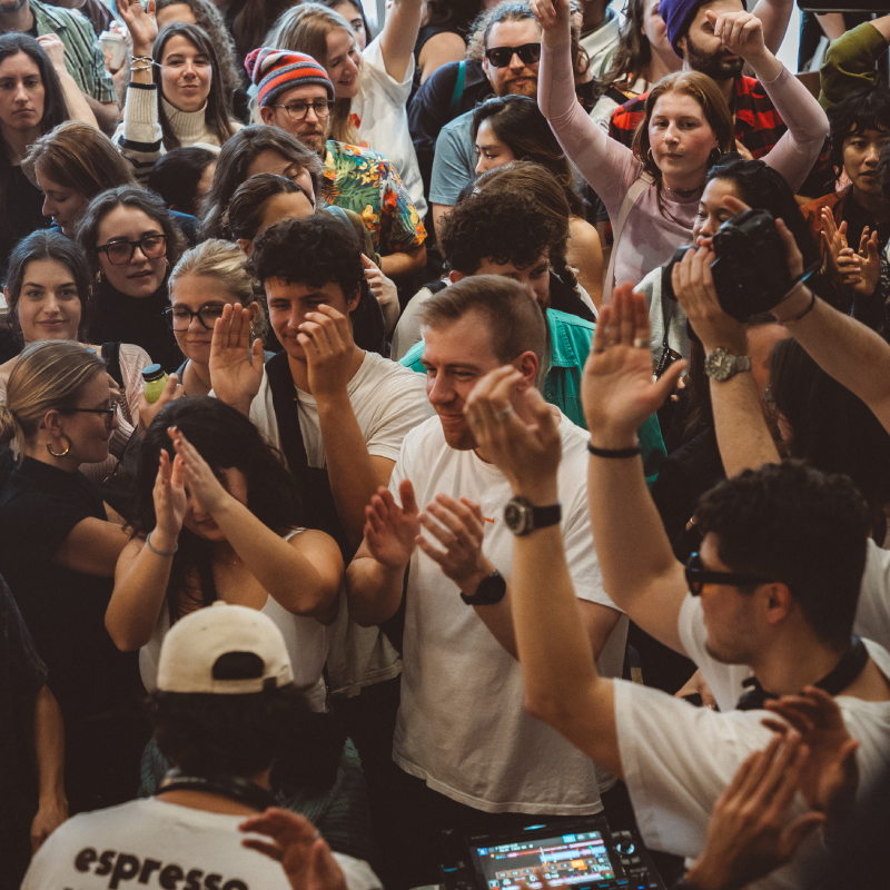 A lively crowd of diverse people gathered closely, many smiling, clapping, and raising hands in celebration or enjoyment.