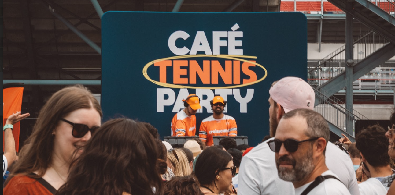 Crowd enjoying an outdoor event in front of a stage with two DJs wearing orange shirts and caps, and a backdrop reading 'CAFÉ TENNIS PARTY'.