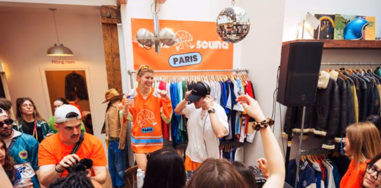 People socializing and a DJ playing music inside a clothing store with colorful shirts and jackets on racks.