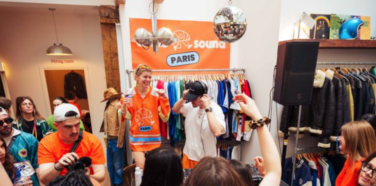 People socializing and a DJ playing music inside a clothing store with colorful shirts and jackets on racks.
