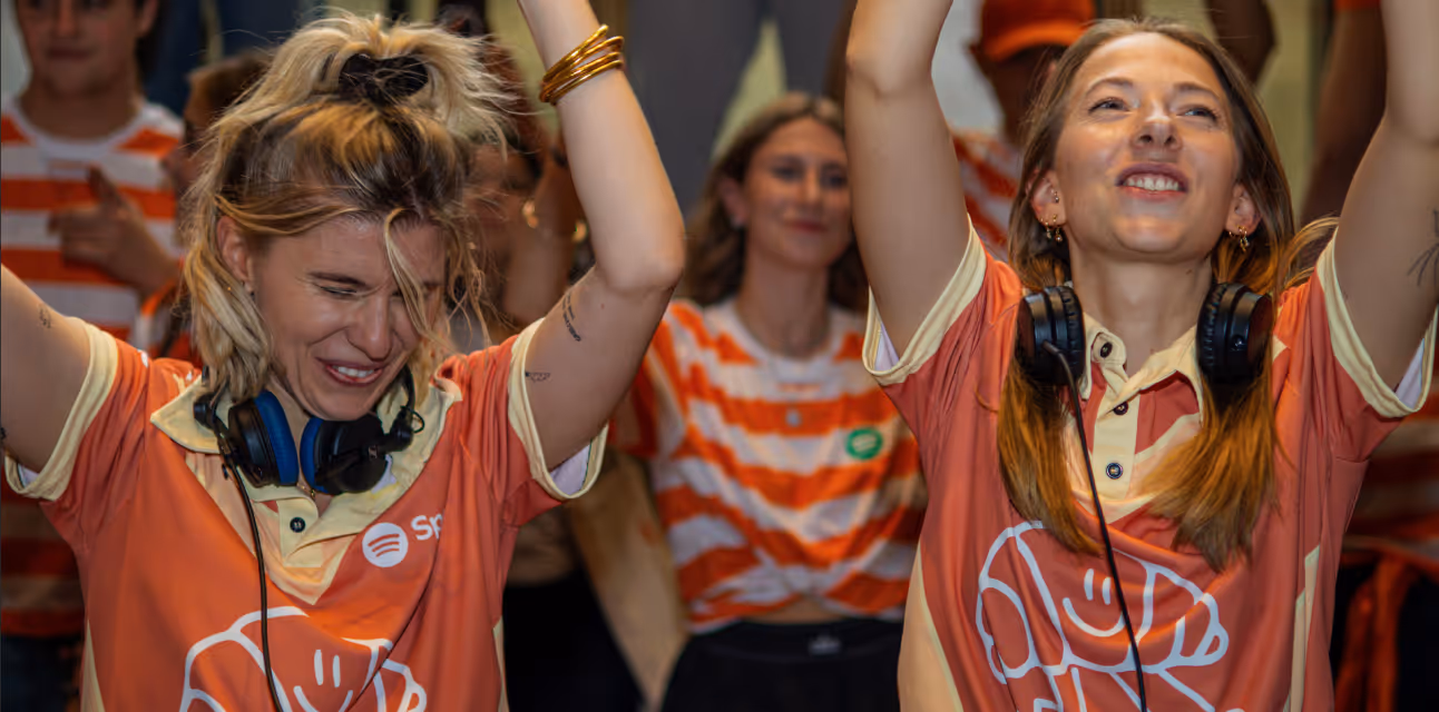 Two women wearing orange Spotify event shirts and headphones joyfully raising their arms at a lively event.