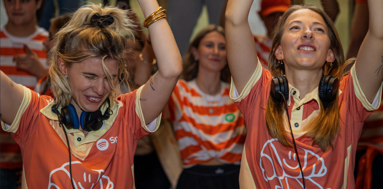 Two women wearing orange Spotify event shirts and headphones joyfully raising their arms at a lively event.