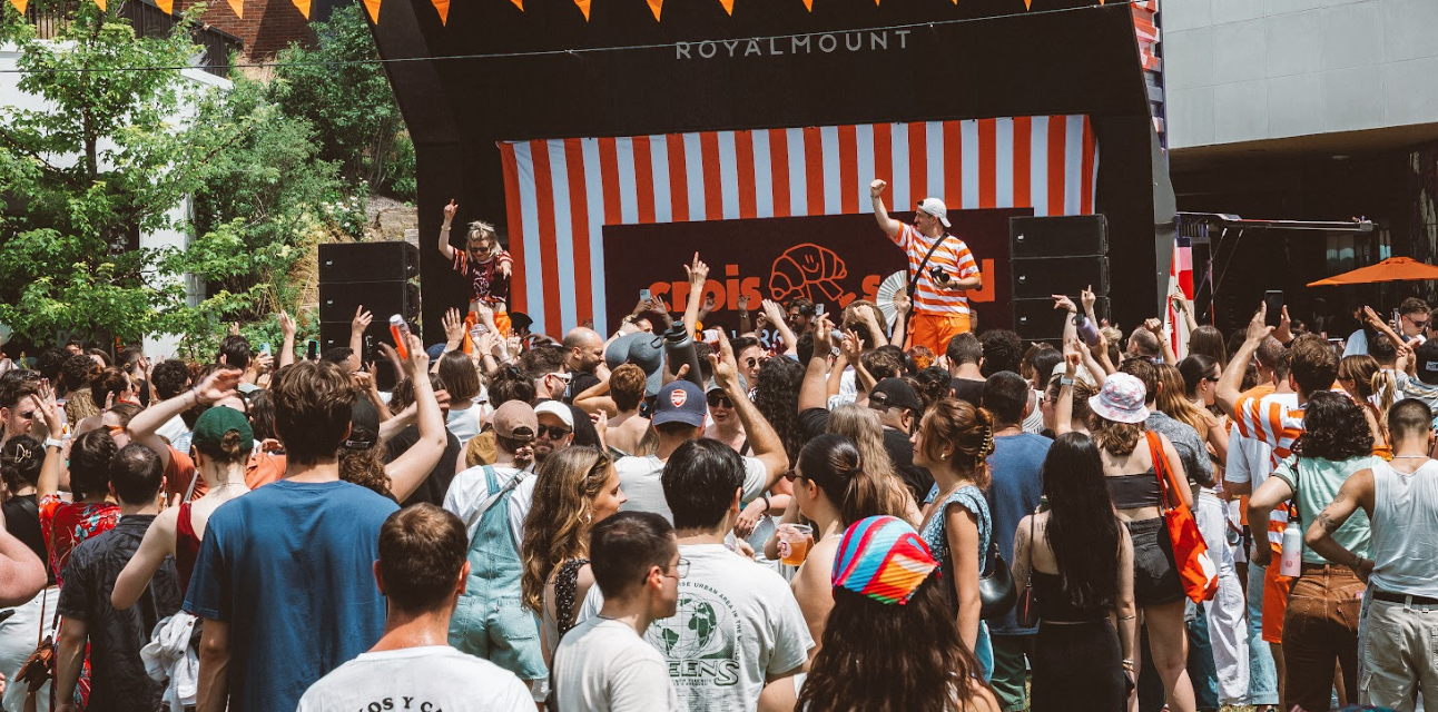 Large crowd enjoying an outdoor music event in front of a stage with red and white striped backdrop at Royalmount.