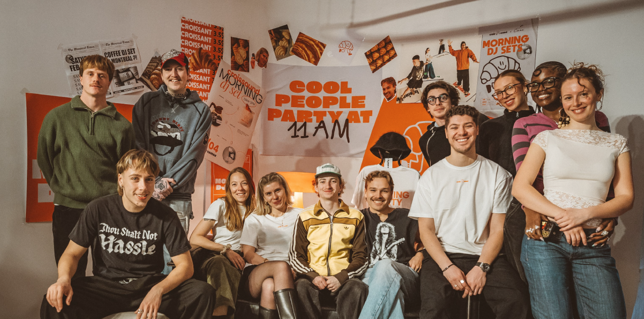 Group of twelve young people smiling and posing indoors in front of a wall covered with orange and white party posters reading 'Cool People Party at 11 AM'.