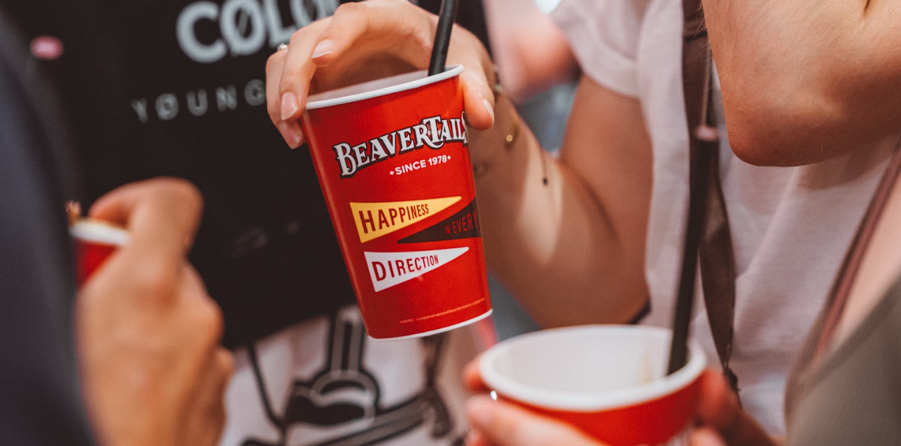 Two people holding red BeaverTails cups with straws, one cup showing the text 'Happiness in every direction'.