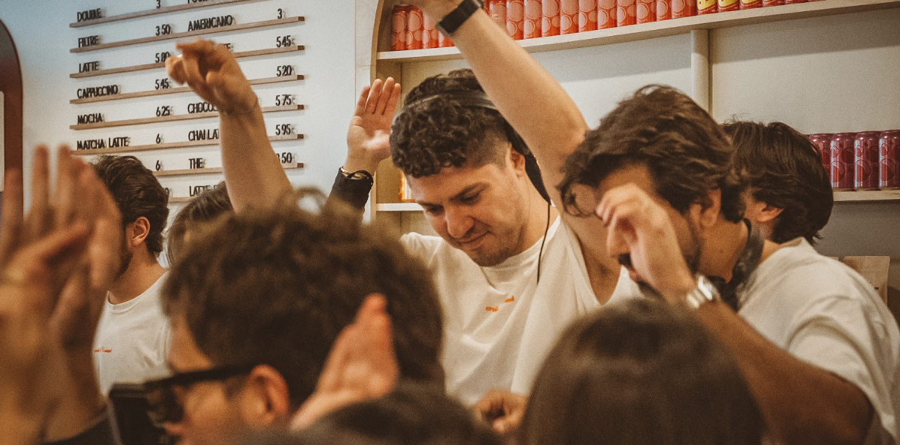 Group of young men dancing and raising their hands in a cozy cafe with drink menu and shelves with cans in the background.