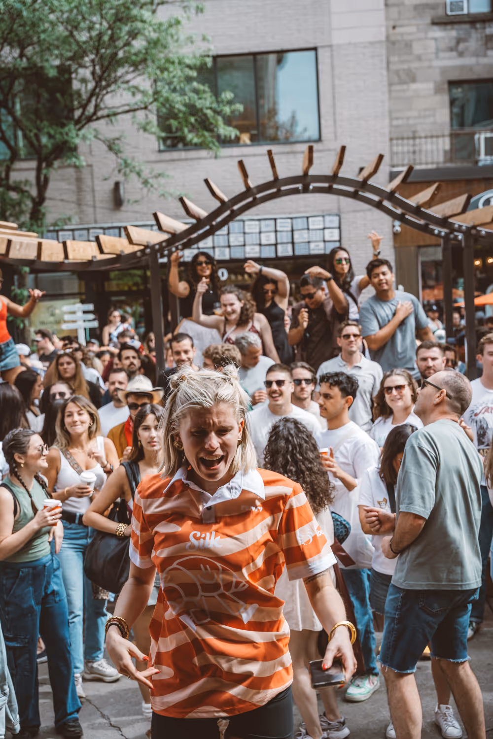 Crowd of people socializing and dancing outdoors under a wooden pergola in an urban setting.