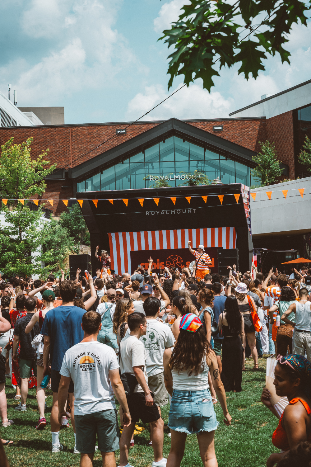Crowd of people enjoying an outdoor event in front of a stage with orange and white stripes at Royalmount complex.