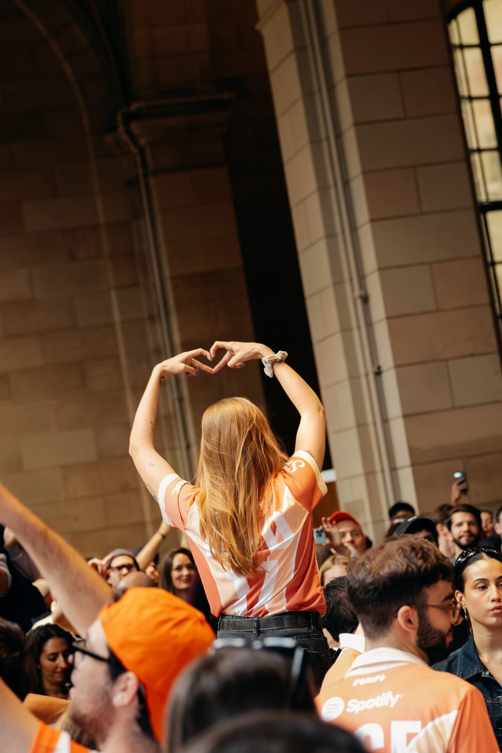 Person with long hair wearing an orange and white Spotify shirt forming a heart shape with their hands above their head in a crowded indoor space.