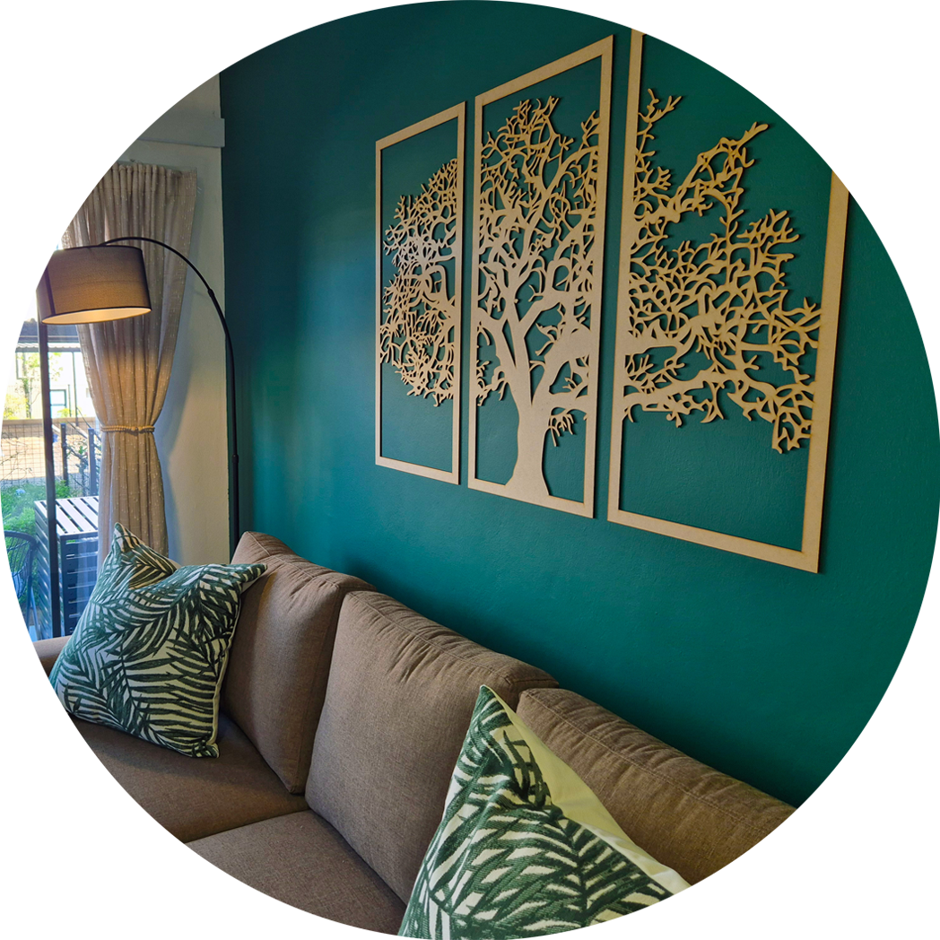 Living room with teal wall featuring wooden tree art in three panels above a brown sofa with green leaf-patterned pillows and a floor lamp near a curtained window.