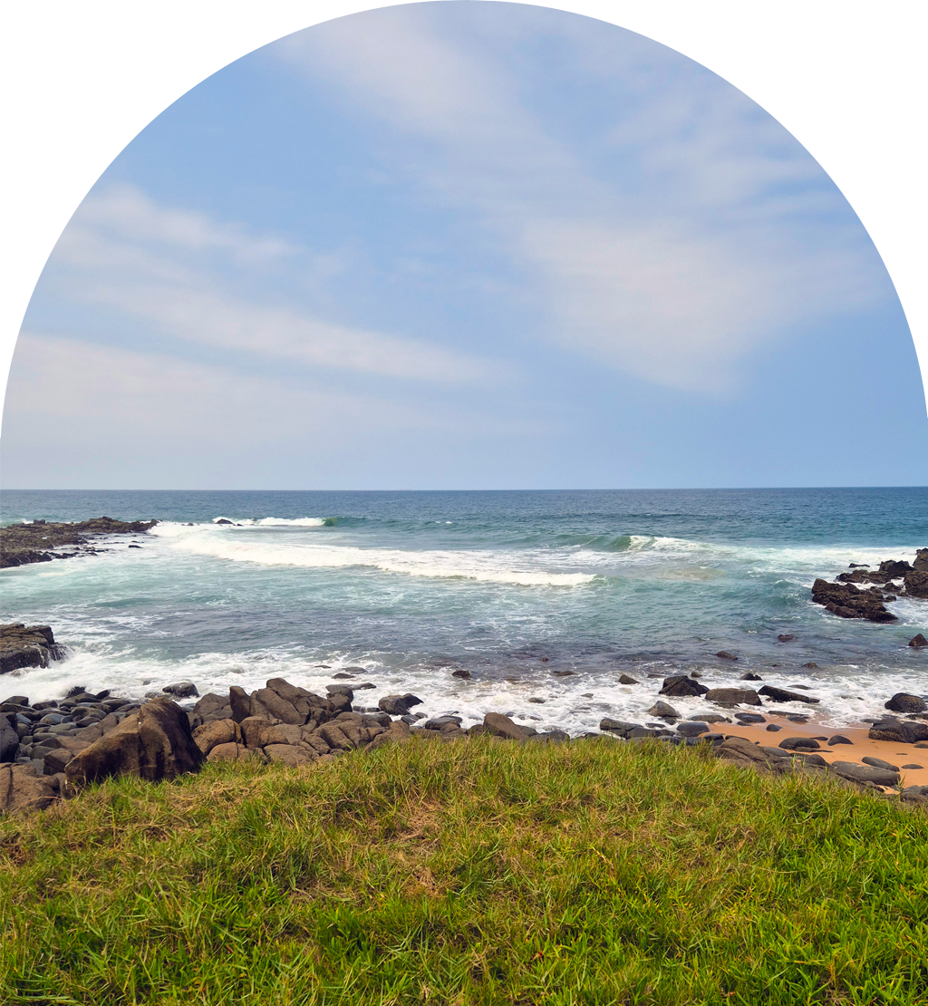 Grassy cliff overlooks rocky shoreline with waves crashing under a partly cloudy sky.
