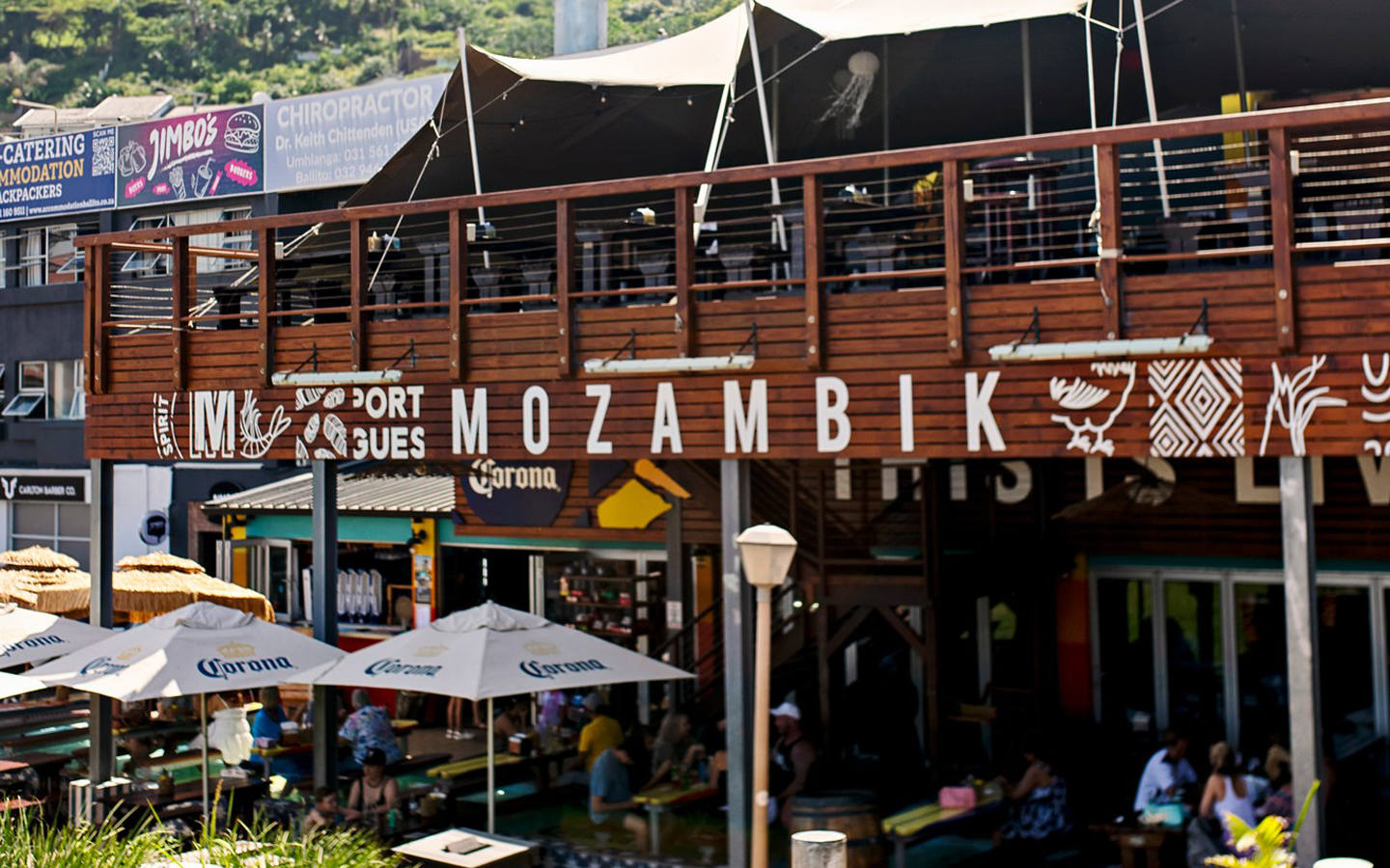 Outdoor seating area of a restaurant named Mozambik with wooden balcony, Corona-branded umbrellas, and patrons dining.