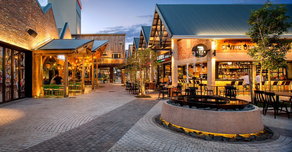 Outdoor shopping center at dusk with lit storefronts, seating areas, and a central fire pit surrounded by trees.