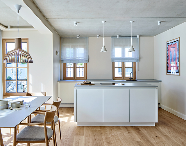 Modern kitchen with a white island, wooden dining table with chairs, pendant lights, two windows with shades, and a framed abstract artwork on the wall.