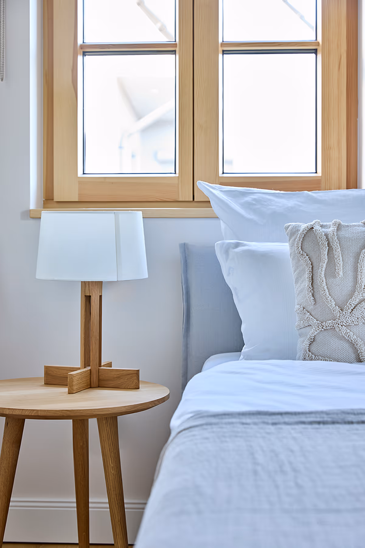 Minimalist bedroom corner with wooden round side table holding a white lamp and a bed with white and textured pillows.