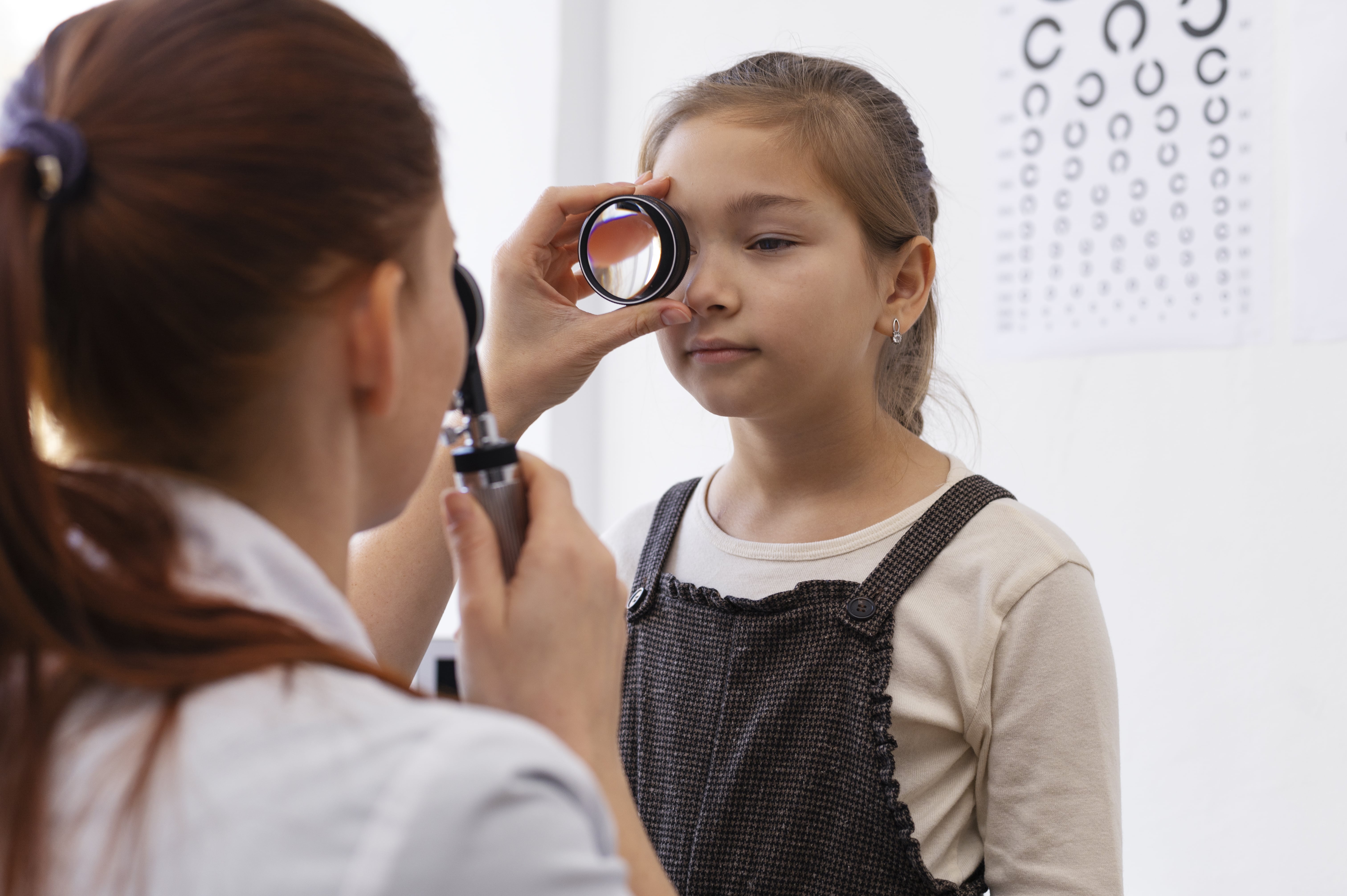 Optometrist examining child's eye with lens during vision test near eye chart