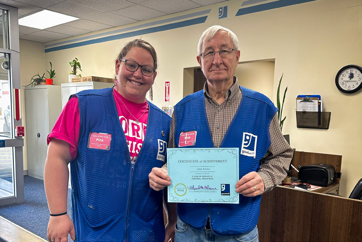 Two Goodwill employees wearing blue vests, one holding a certificate of achievement.