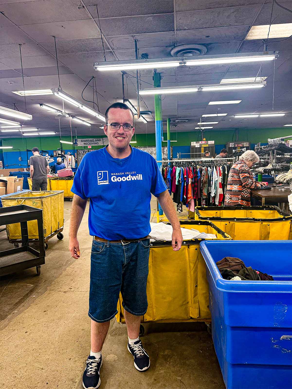 Smiling man wearing glasses and a blue Wabash Valley Goodwill t-shirt standing inside a thrift store with clothing racks and yellow bins around.