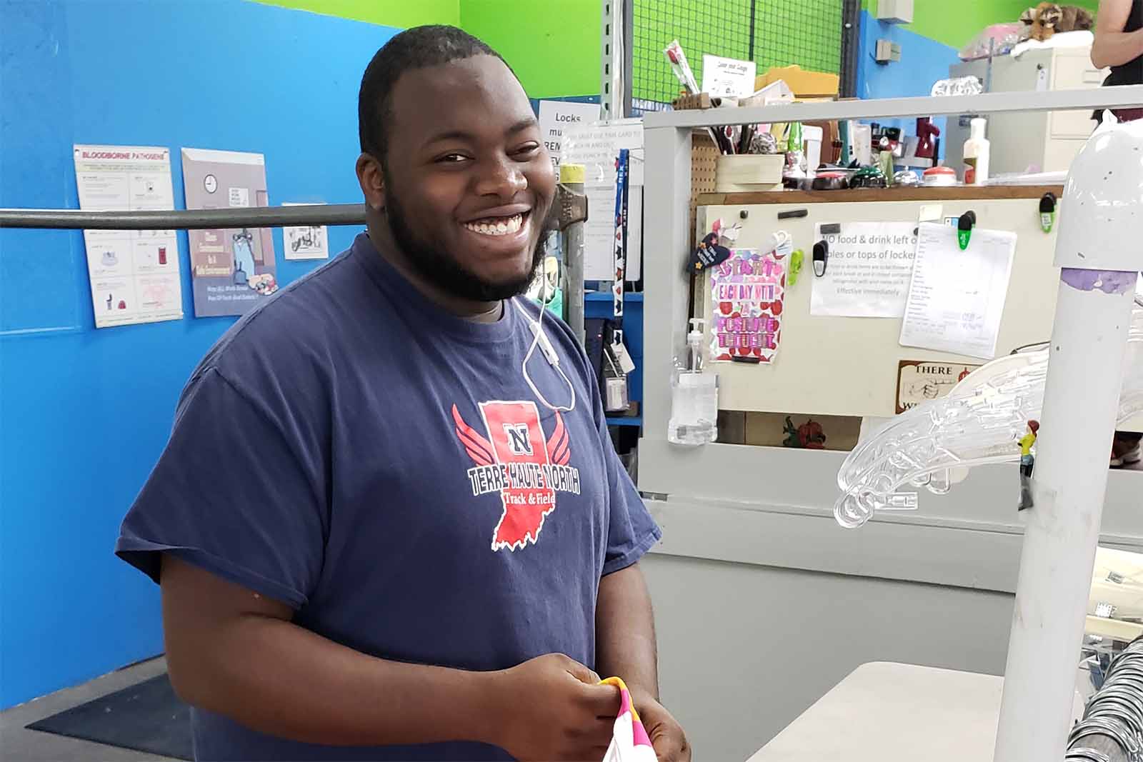 Smiling man wearing a Terre Haute North Track & Field t-shirt standing indoors near a counter with various items on shelves behind him.