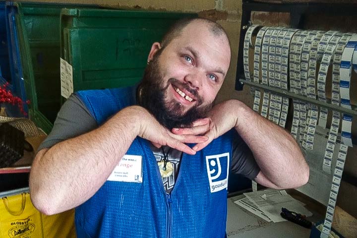 Smiling man with a beard wearing a blue Goodwill vest posing with hands under chin in a workspace with bins and label rolls.