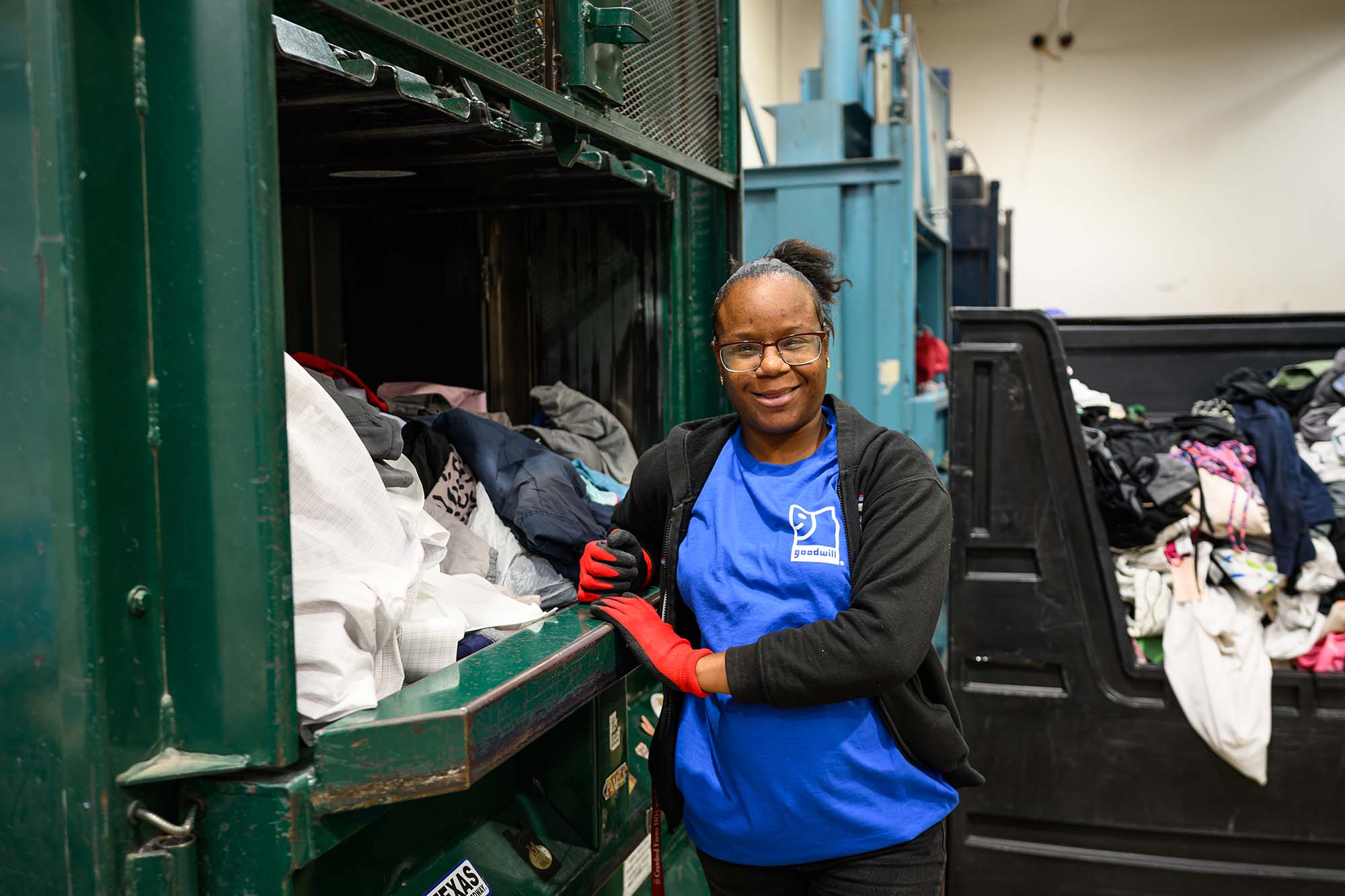 Smiling woman wearing red gloves and a blue Goodwill shirt sorting clothes by a large green recycling bin filled with garments.