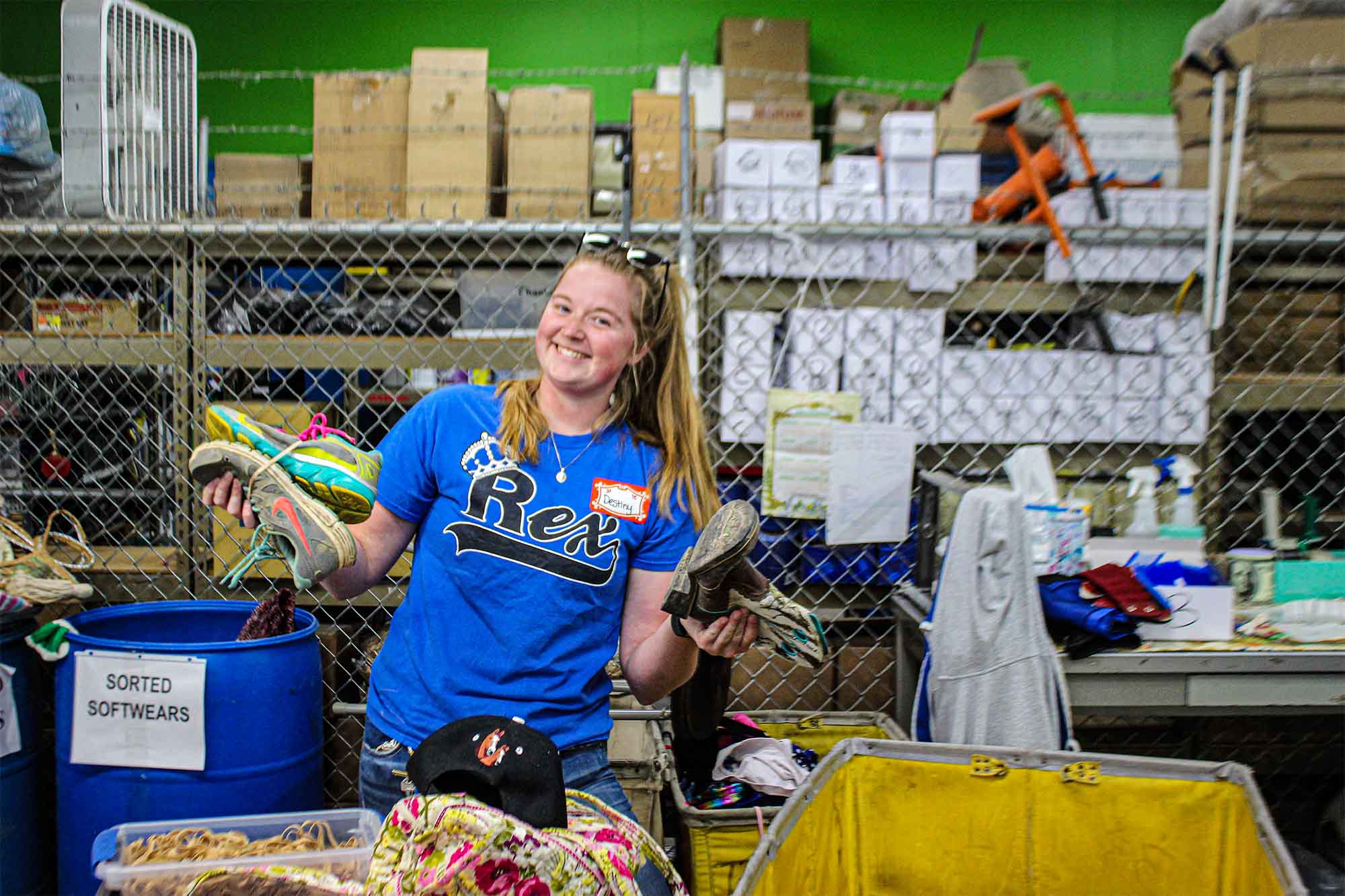 Smiling female Goodwill employee in a blue t-shirt holding pairs of shoes inside a sorting warehouse with containers and boxes behind her.