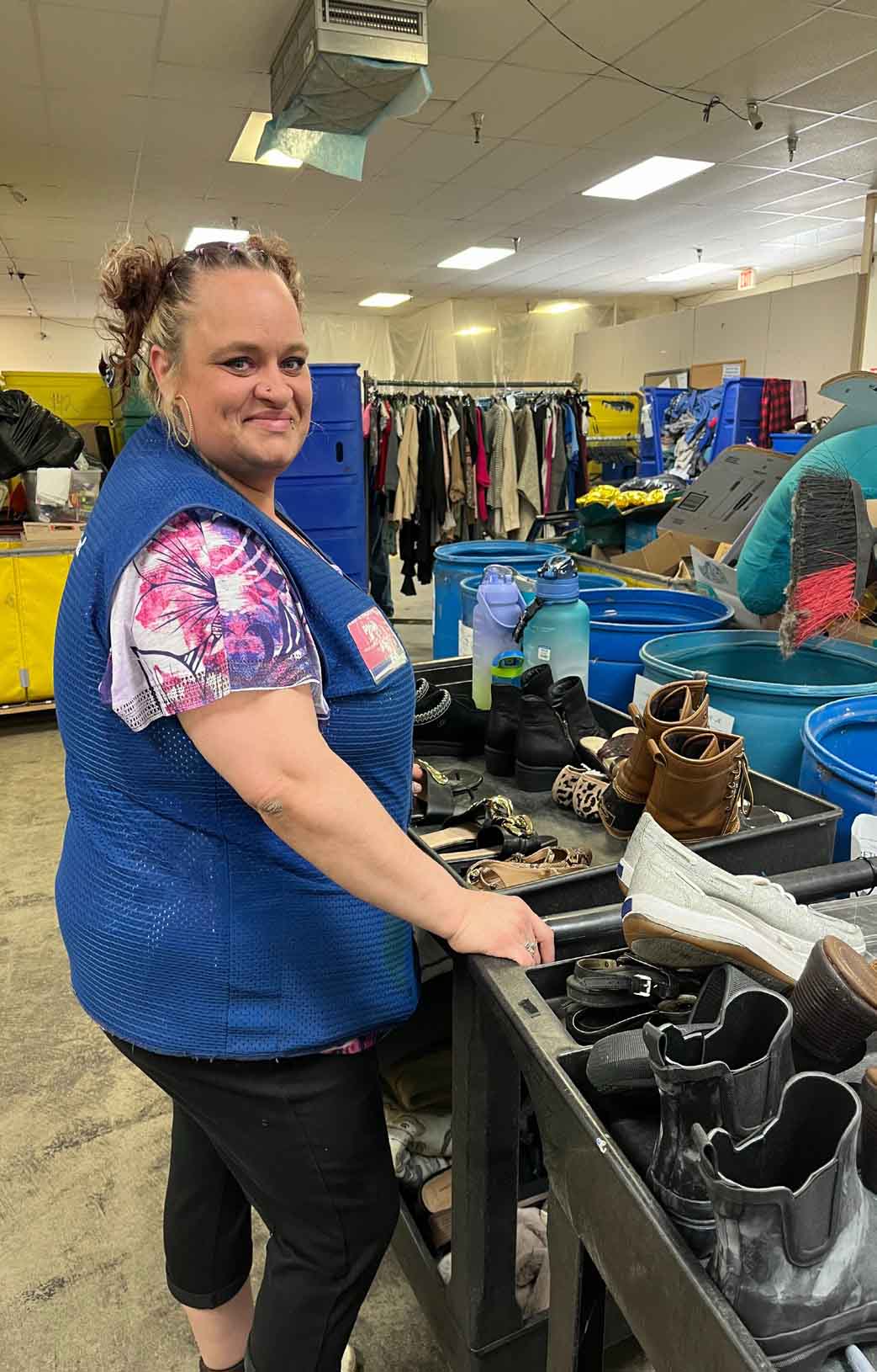 Woman in a blue Goodwill vest standing beside bins filled with shoes in a warehouse-like setting with clothing racks in the background.