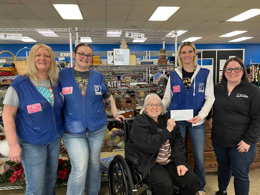 Five women posing inside a thrift store, two wearing blue Goodwill vests, one seated in a wheelchair holding a document.