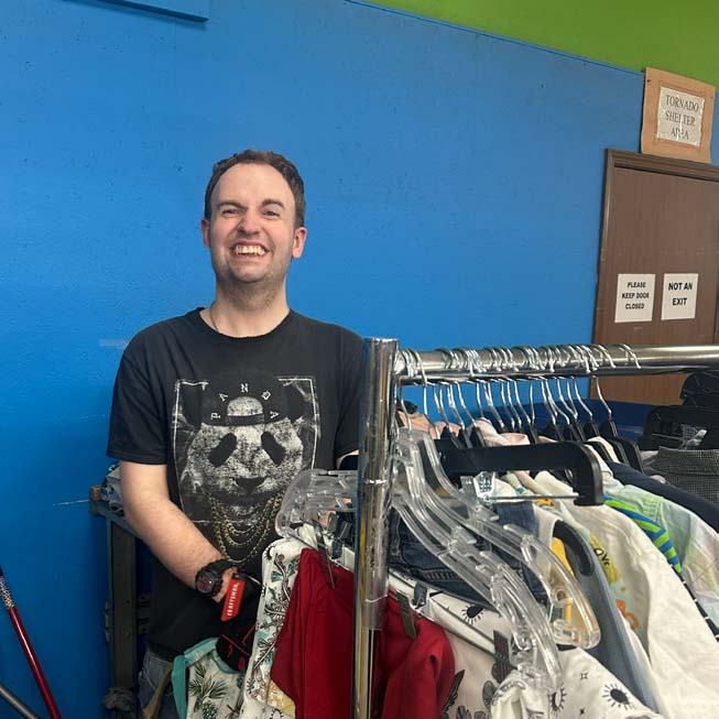 Smiling Goodwill team member standing behind a clothing rack with various clothes on hangers against a blue and green wall.