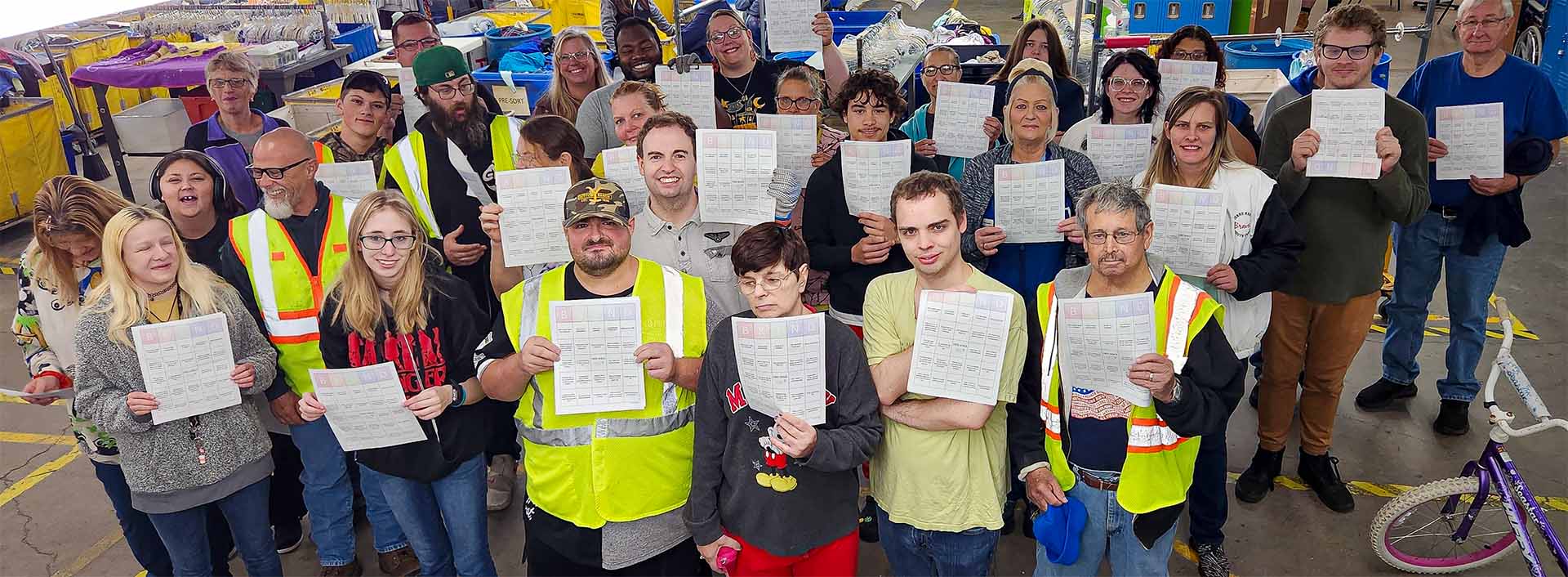 Group of diverse Goodwill production team workers in high-visibility vests holding printed sheets, standing in an industrial workspace.