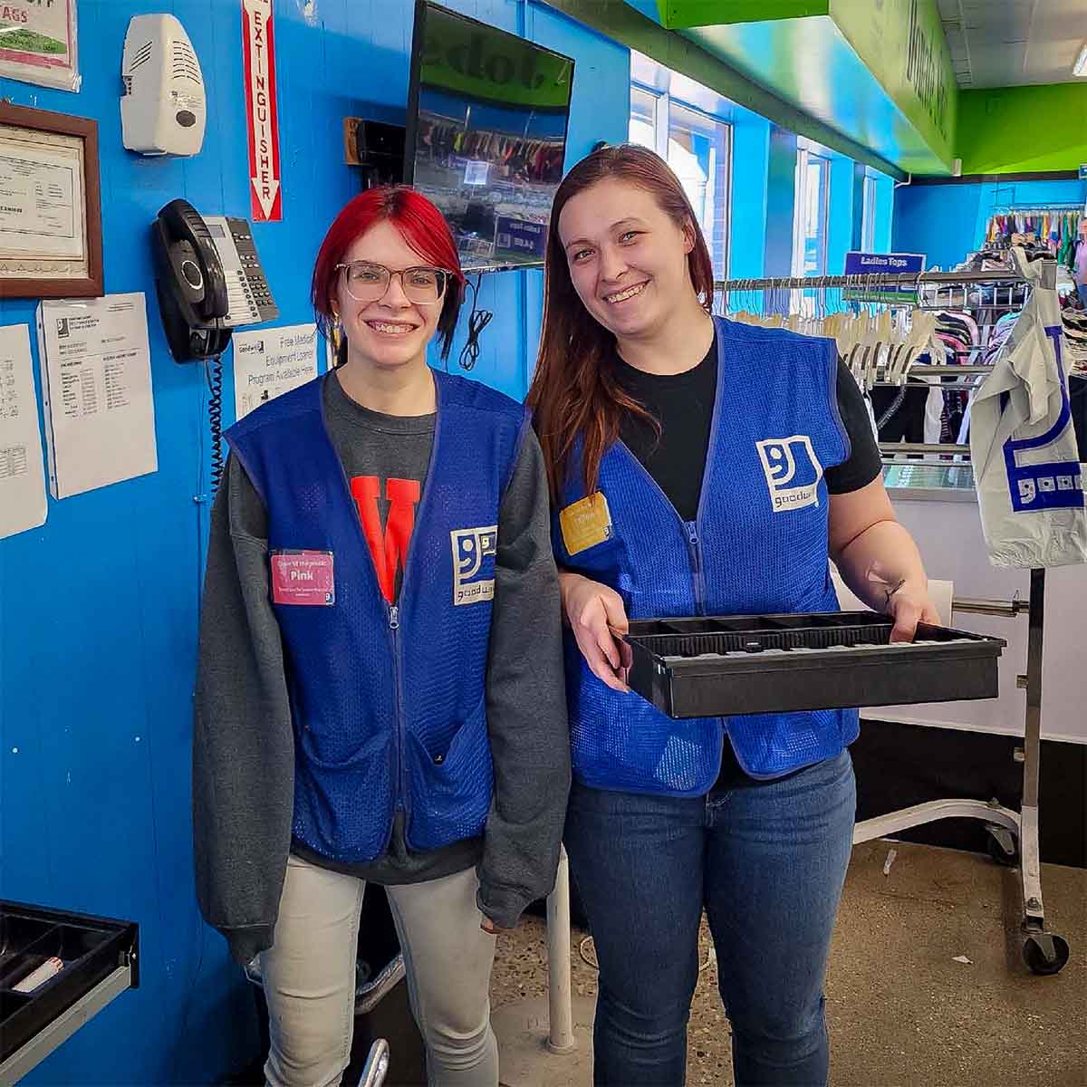 Two smiling Goodwill employees wearing blue vests standing inside a store with clothing racks in the background.