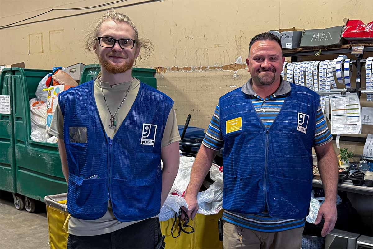 Two Goodwill employees wearing blue vests standing in a storage area with bins and supplies behind them.