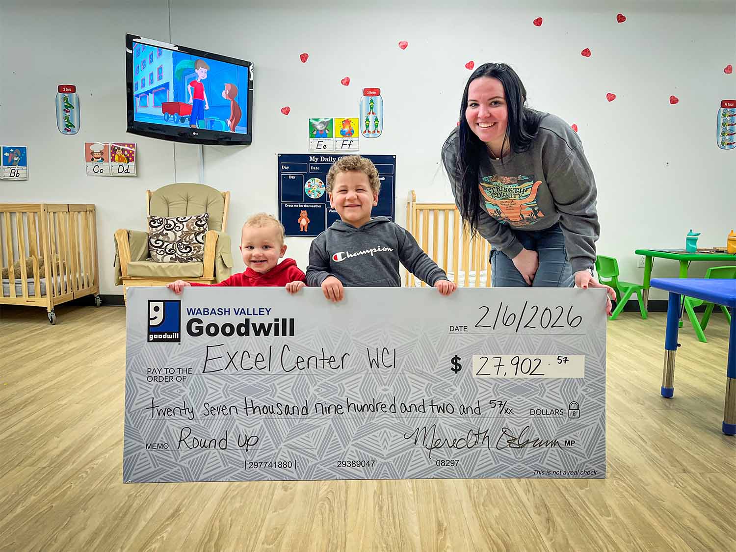 Two children and a woman smiling and holding a large ceremonial check from Wabash Valley Goodwill to Excel Center WCI for $27,902.57 in a classroom.