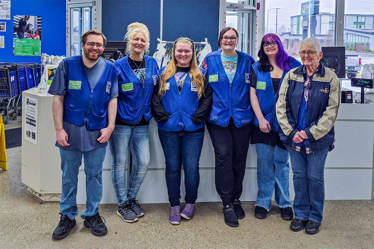 Group of six Goodwill retail employees wearing blue vests standing inside a store with a counter and shopping carts in the background.