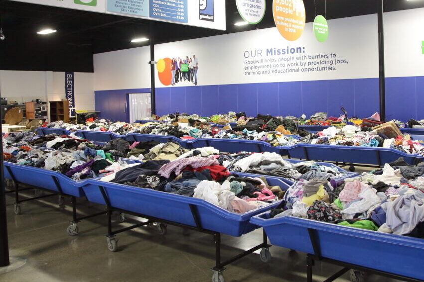 Rows of blue bins filled with various clothes inside a Goodwill thrift store donation area under a mission statement wall.