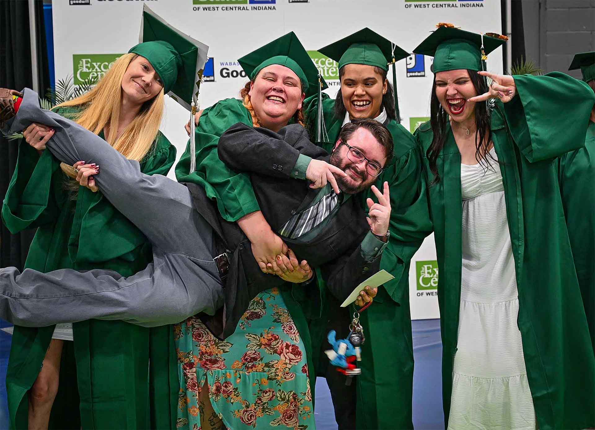 Four Excel Center graduates in green caps and gowns posing playfully while holding a smiling man in a suit horizontally.