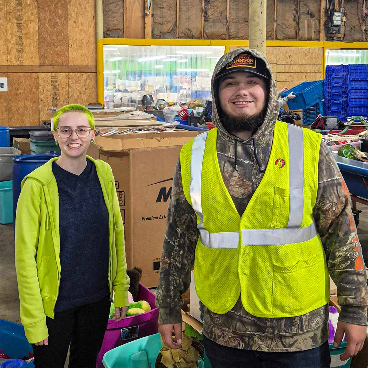 Two smiling Goodwill workers inside a warehouse surrounded by bins and boxes, one wearing a yellow safety vest and camouflage hoodie, the other with bright green hair and glasses.