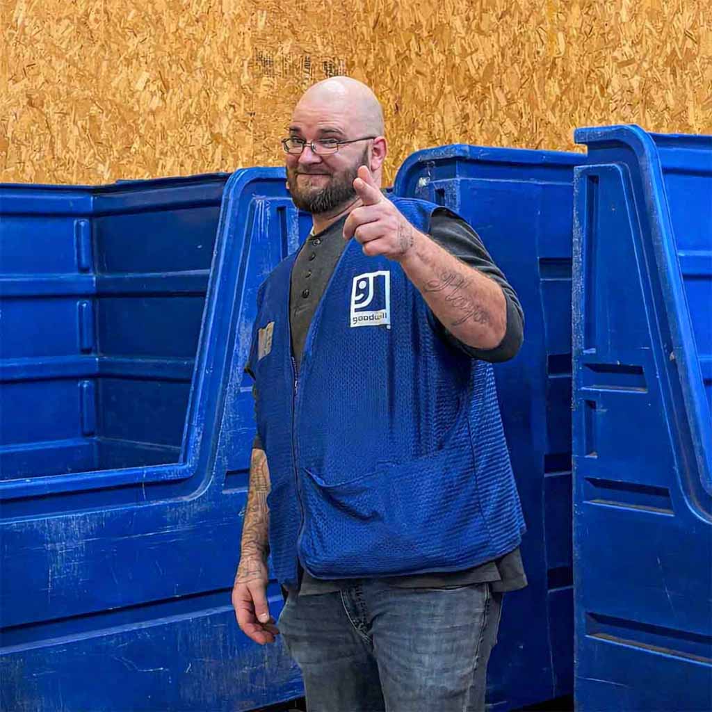 A man with glasses and a beard wearing a blue Goodwill vest standing in front of blue bins and a wooden wall.