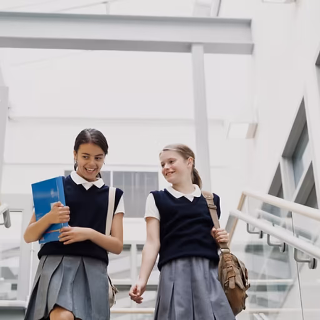 Two female students, one holding a folder and one with a bag over her shoulder walking down a school staircase in school uniform.