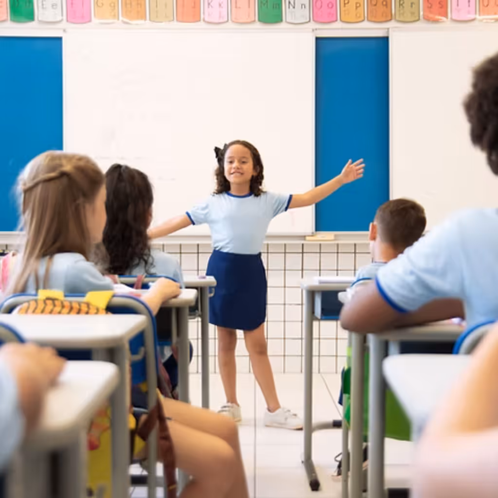 A young female student standing at the front of a classroom presenting and smiling with her arms out wide.