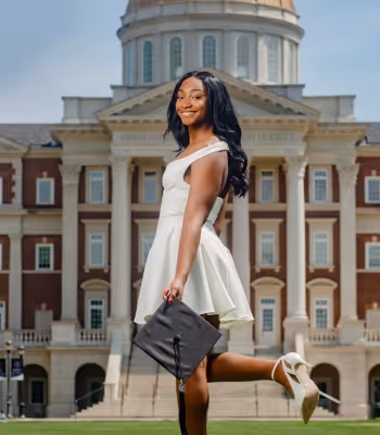 Picture of a black female student in a white dress holding a graduation cap in front of an elegant building.