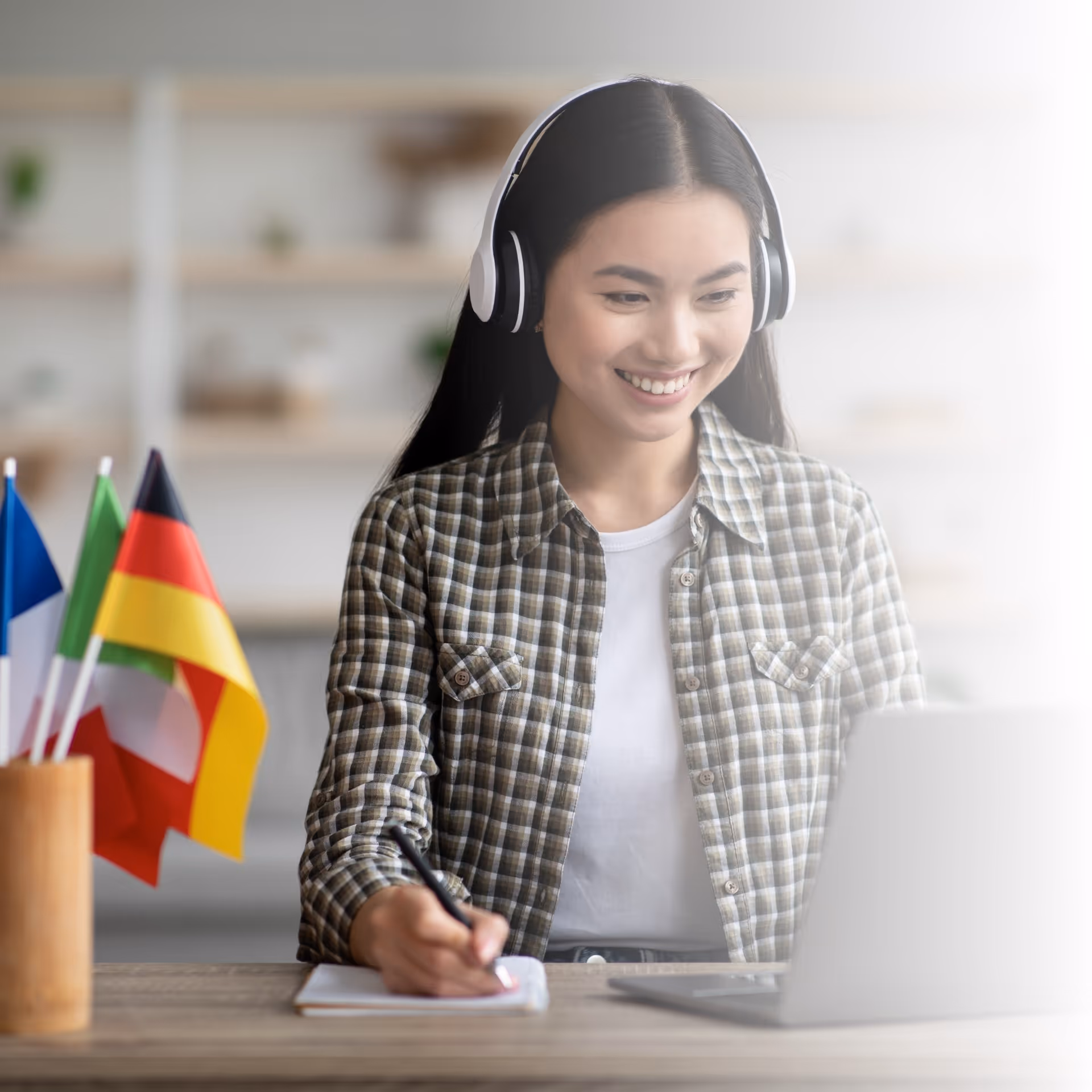 A female asian student with headphones on making notes in front of a laptop with multiple flags of countries to her right.