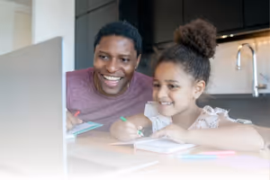 A father and daughter looking at a computer laptop doing homework smiling.