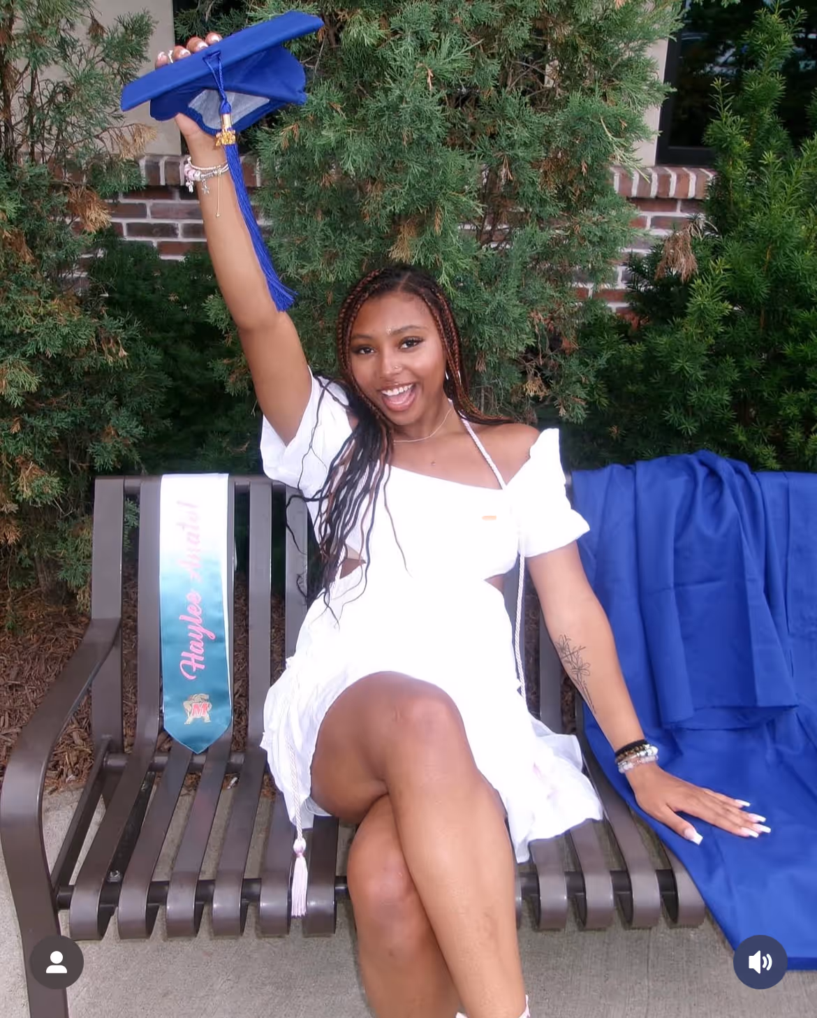 A young female student in a white dress holding a blue graduation cap above her head to celebrate.