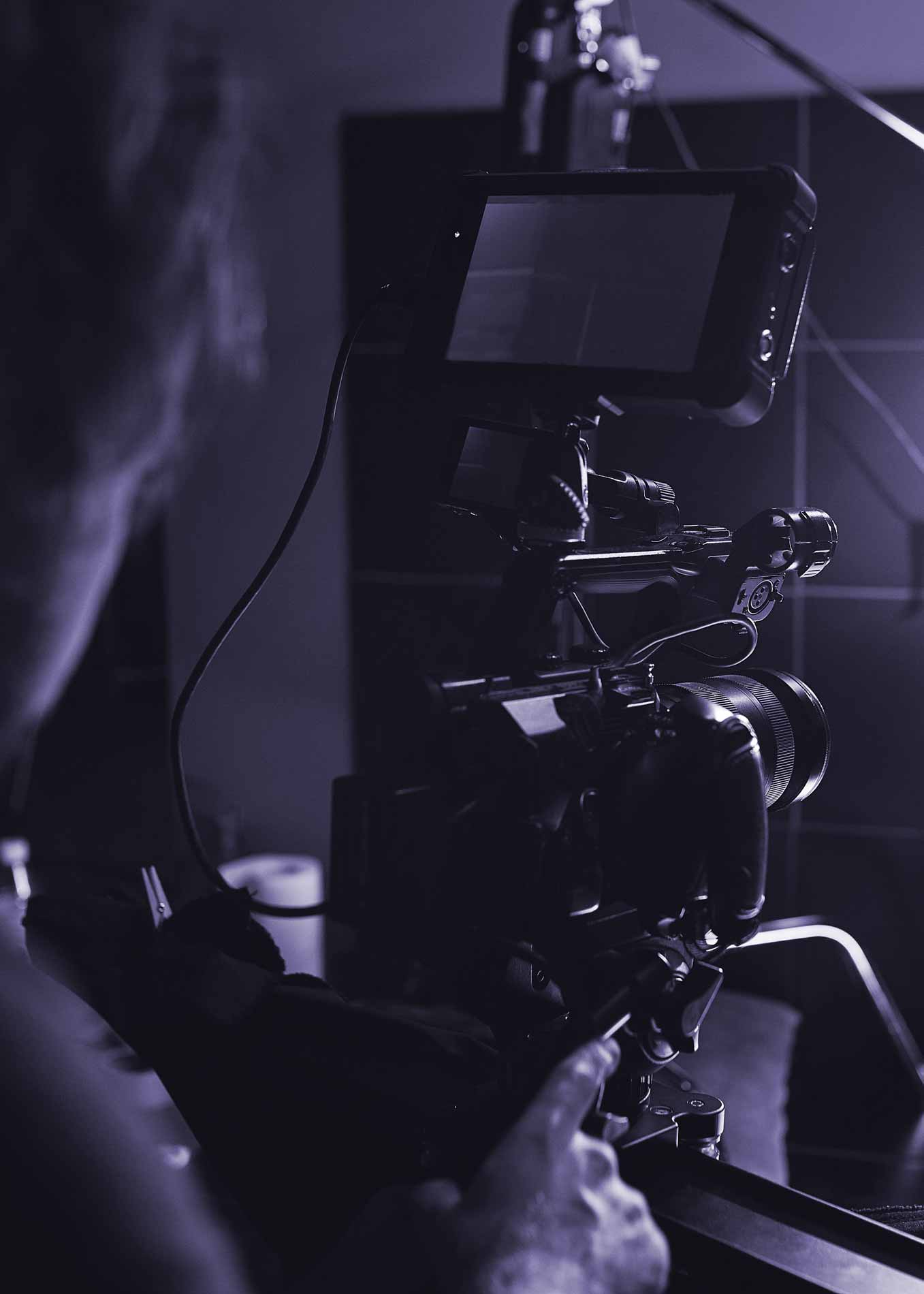 Close-up van een persoon die een professionele videocamera bedient met een monitor en lens in een donkere omgeving.