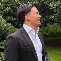 Een man in een zwart pak en witte blouse die buiten lacht met groene struiken op de achtergrond.