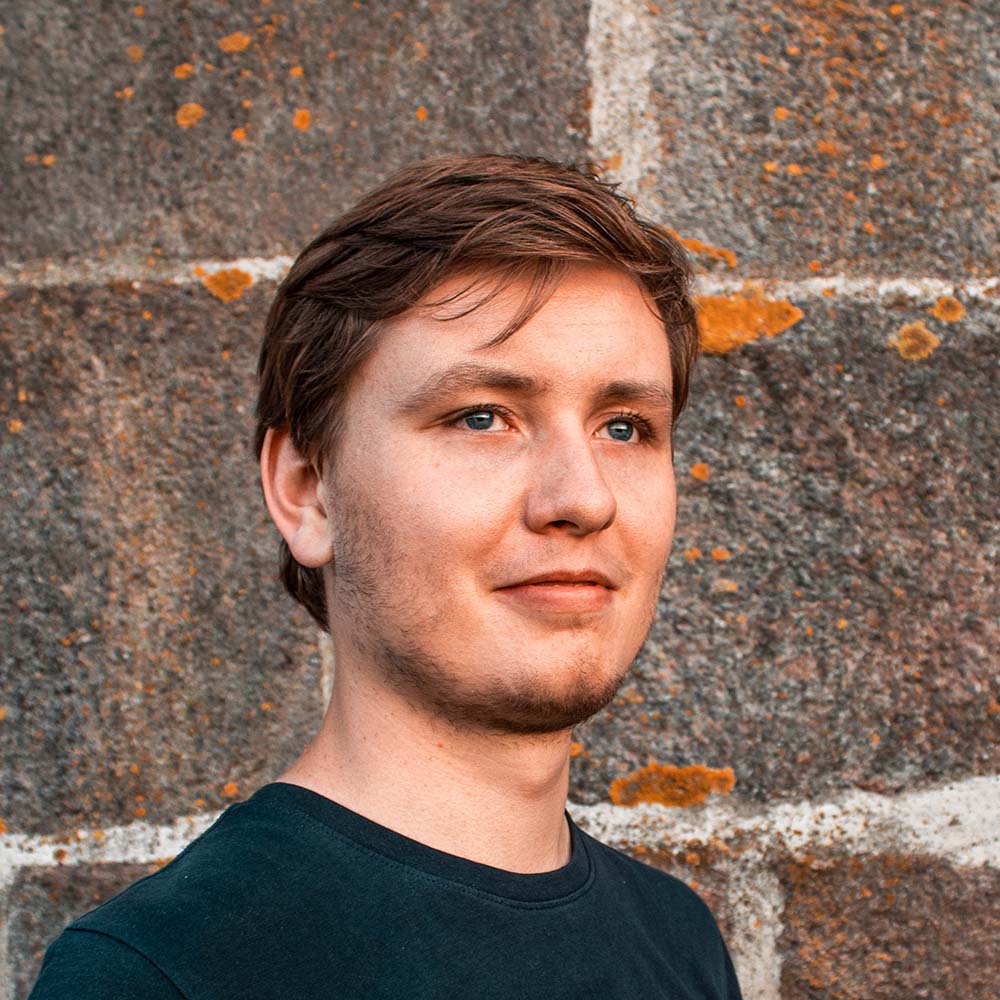 Jonge man met kort bruin haar en blauwe ogen draagt een donker T-shirt voor een stenen muur met oranje mos.