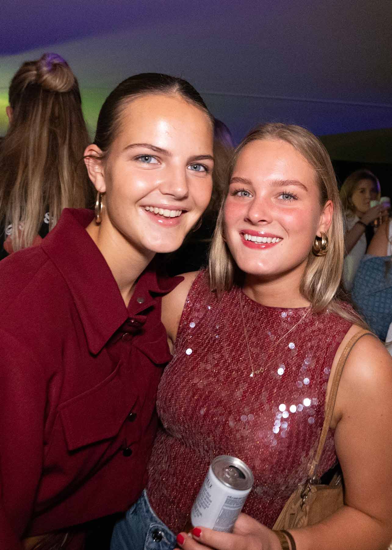 Twee jonge vrouwen glimlachen en poseren samen op een feest, één draagt een bordeauxrode blouse en de ander een glinsterende rode top met een drankje in haar hand.