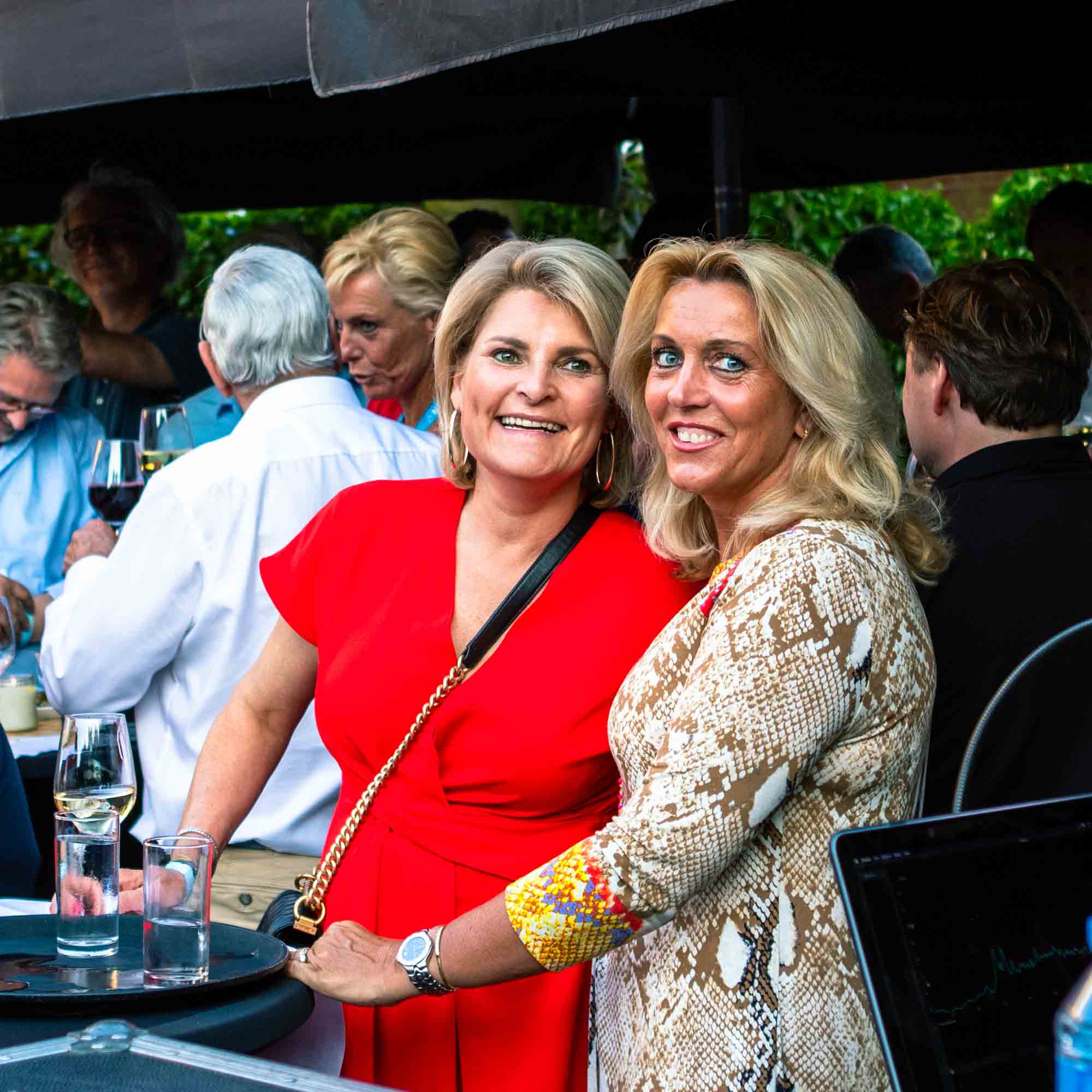 Twee vrouwen lachen samen buiten bij een sociale bijeenkomst, één draagt een rode jurk en de ander een slangenhuidpatroon blouse.